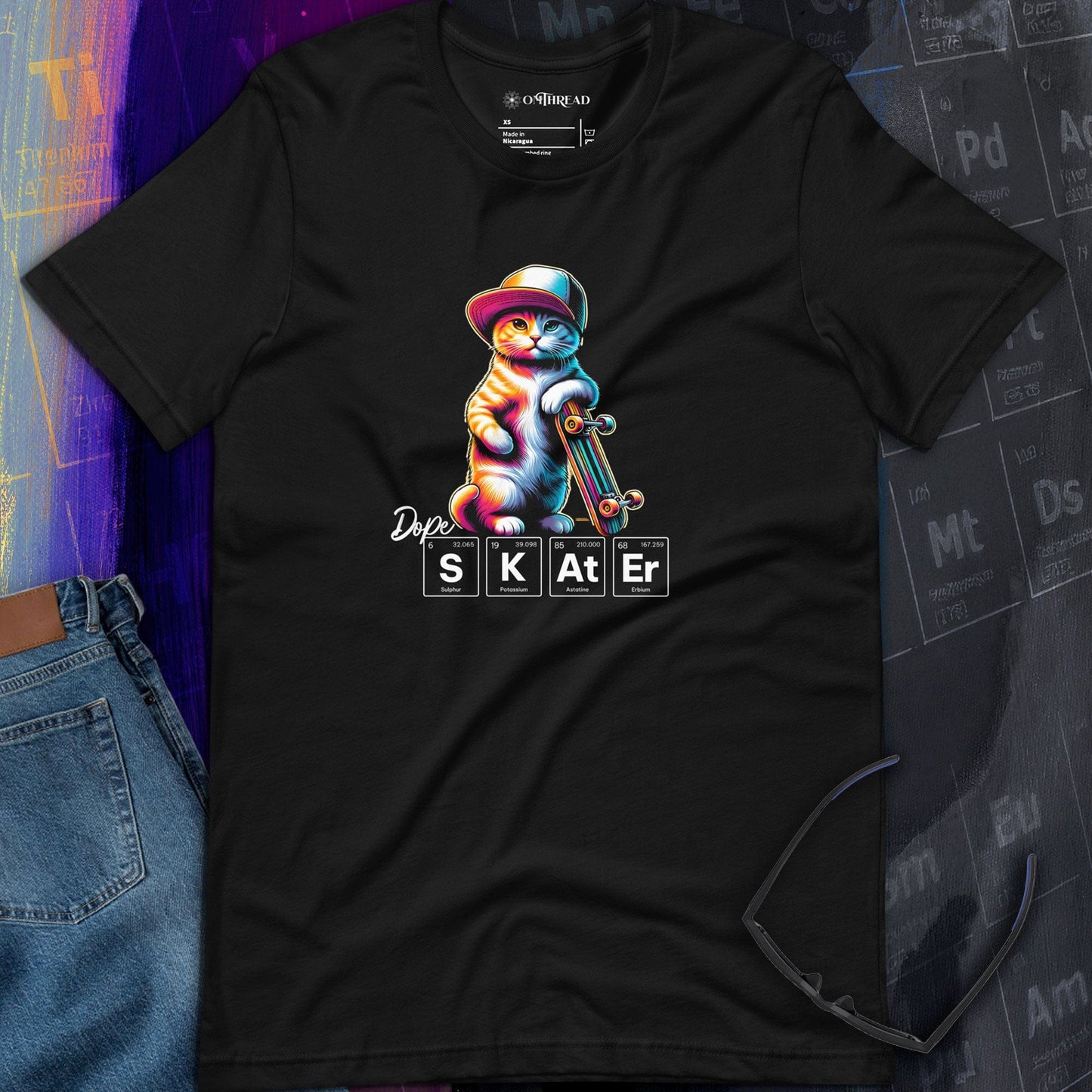 OMTHREAD black t-shirt featuring a colorful illustration of a cat wearing a hat and holding a skateboard. Below the cat, the word "SKATER" is spelled out using periodic table elements: sulfur (S), potassium (K), astatine (At), and erbium (Er).