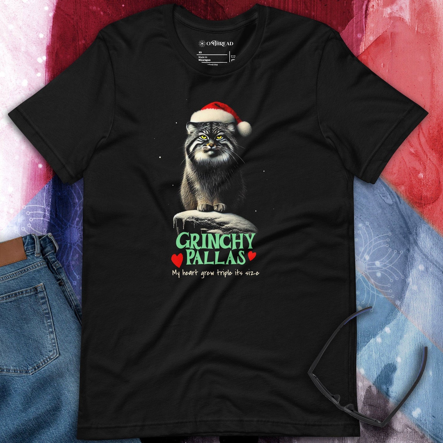OMThread black t-shirt featuring a grumpy Pallas cat sitting on a snowy rock, wearing a Santa hat. The text below reads "GRINCHY PALLAS" in green and red, followed by the phrase "My heart grew triple its size" in a handwritten style.