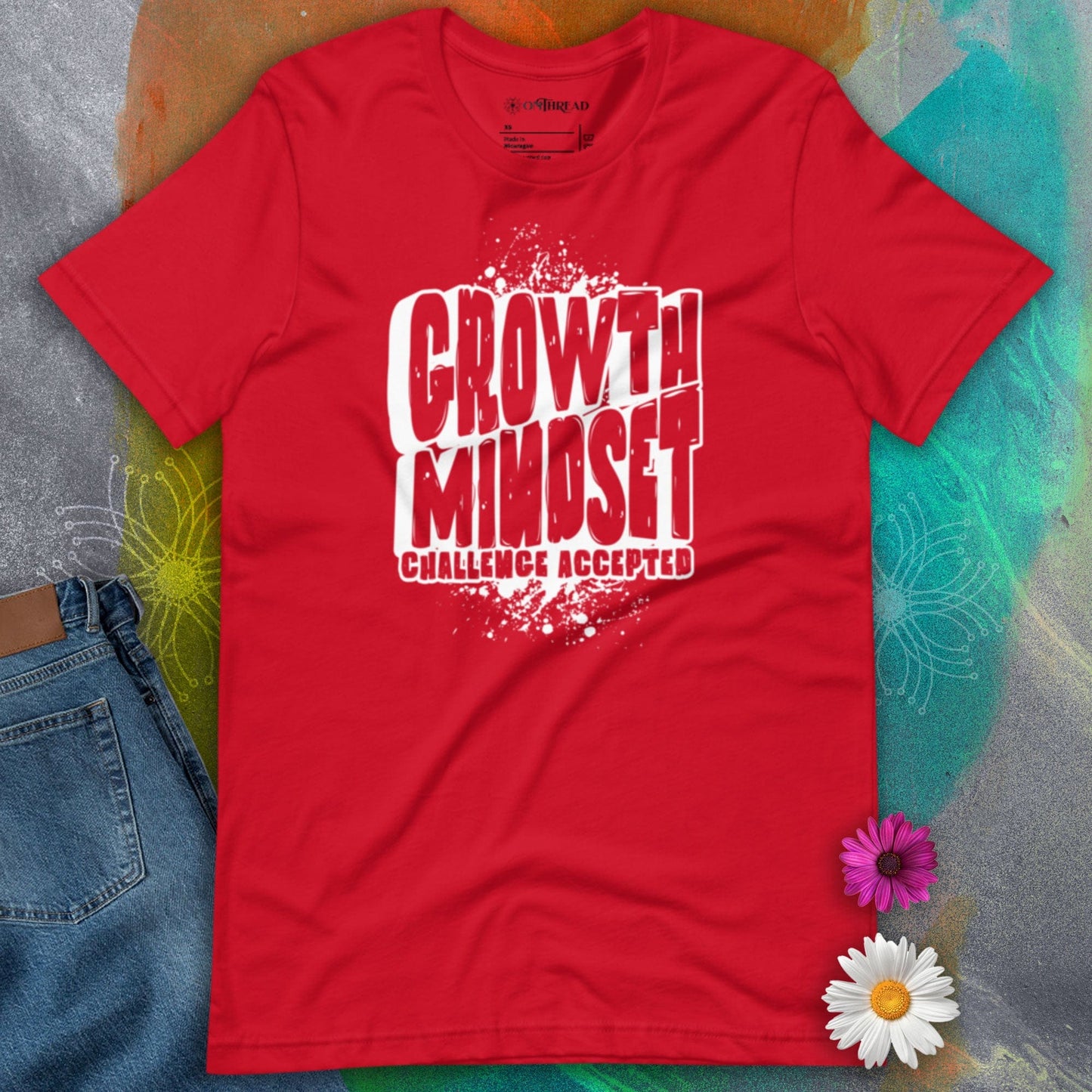 OMTHREAD red t-shirt with bold white text reading "Growth Mindset Challenge Accepted" in a distressed font, laid flat on a colorful background with flowers and denim.