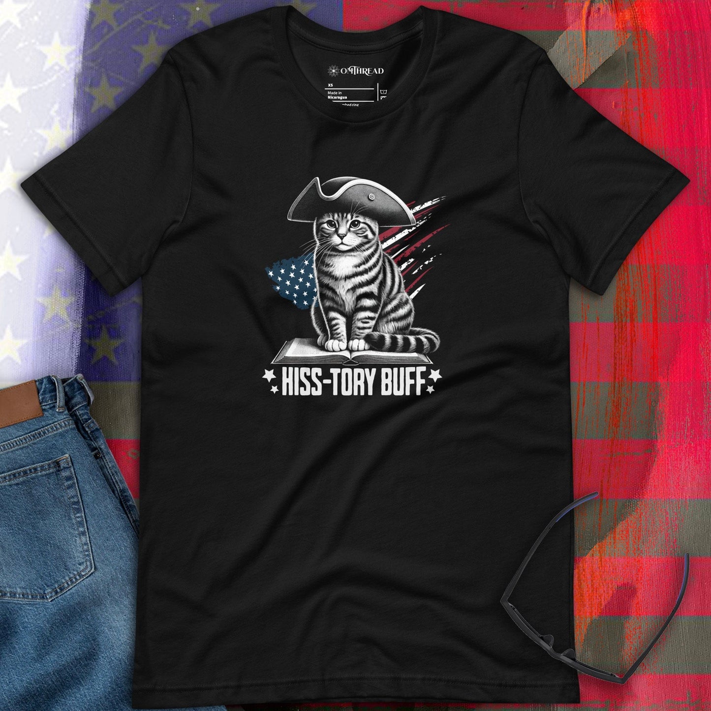 OMThread Hiss-tory Buff Cat T-Shirt featuring a colonial-style tabby cat sitting on a book, wearing a tricorn hat, with an American flag background. The design includes bold text that reads 'Hiss-tory Buff.' Printed on a black unisex t-shirt.