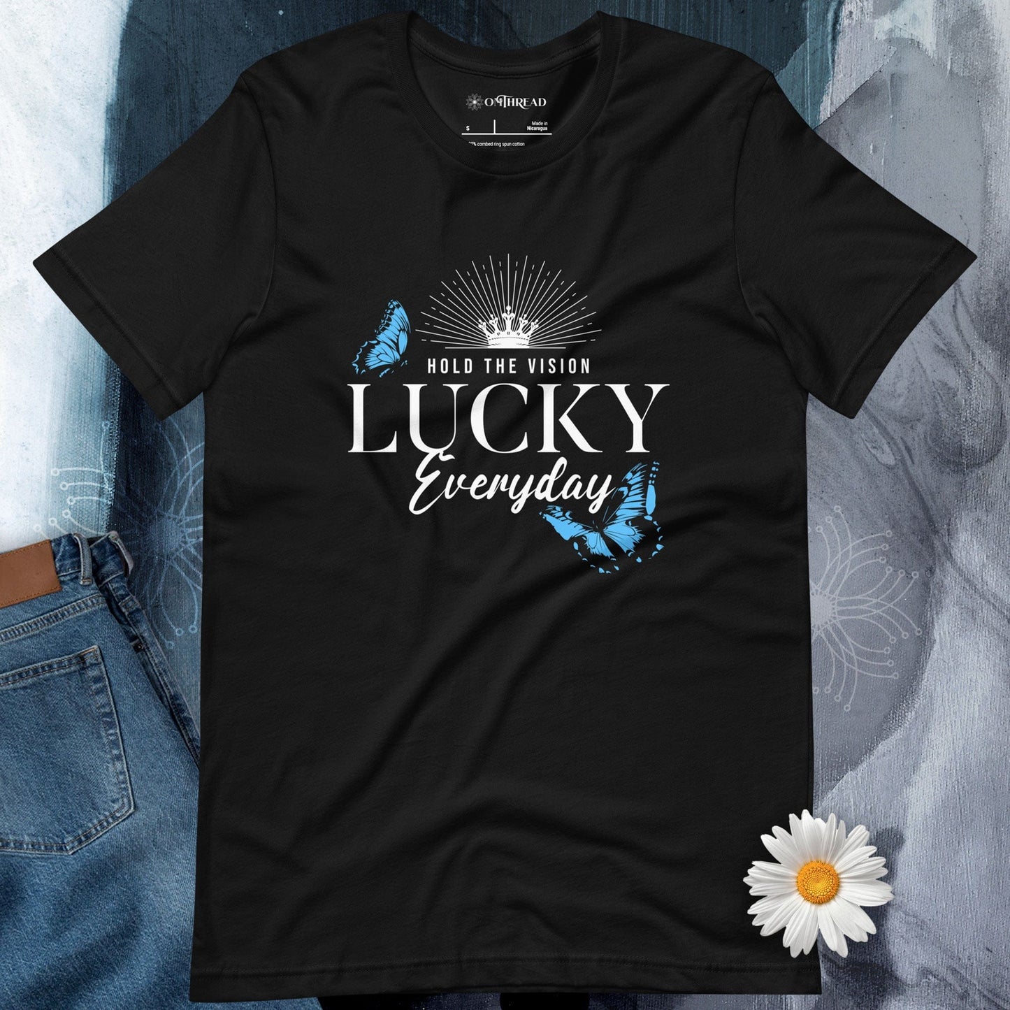 OMTHREAD black t-shirt featuring the text "Hold the Vision" above "LUCKY Everyday" with a crown and butterfly designs in blue, creating a motivational and inspirational visual.
