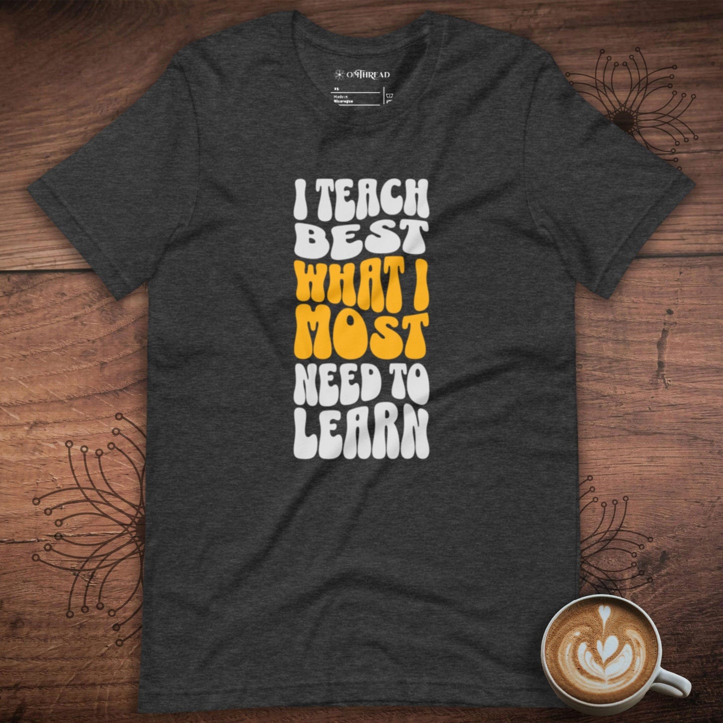 OMTHREAD dark grey heather t-shirt with bold white and yellow text reading "I Teach Best What I Most Need to Learn" in a retro-style font.