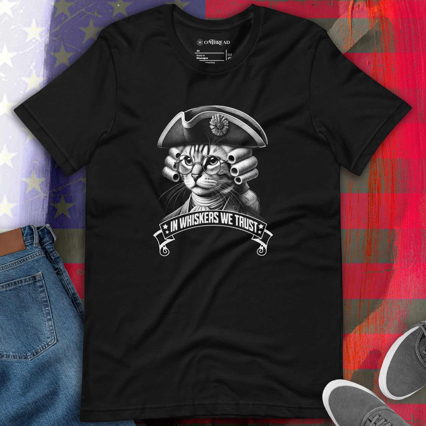 OMTHREAD black t-shirt featuring a detailed cat illustration dressed in colonial attire, wearing a tricorn hat, glasses, and a powdered wig. The design includes the phrase 'In Whiskers We Trust' in a bold banner.