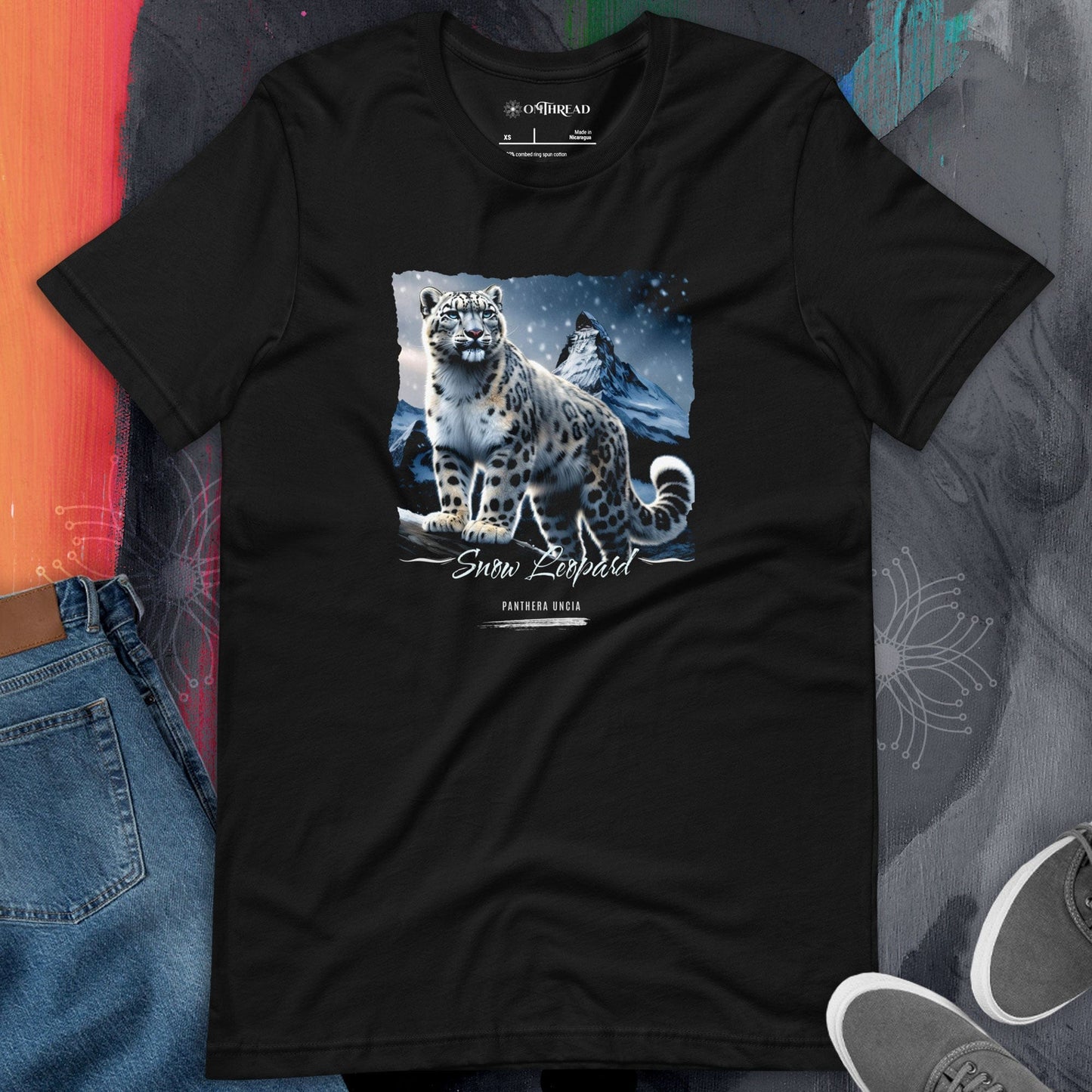 OMThread black t-shirt featuring a detailed illustration of a snow leopard standing on a rocky ledge with a mountain landscape in the background. The text "Snow Leopard" and "Panthera Uncia" is printed below the artwork. The design is centered on the shirt, with a realistic and artistic style.