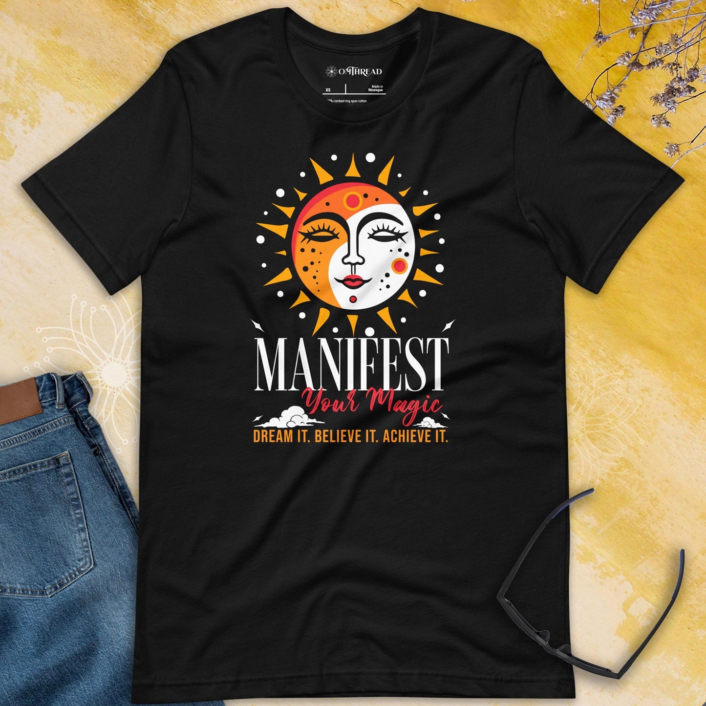 OMTHREAD black t-shirt with a colorful sun design featuring the words "MANIFEST Your Magic" in white and red font, with the phrase "DREAM IT. BELIEVE IT. ACHIEVE IT." in yellow text below the sun.