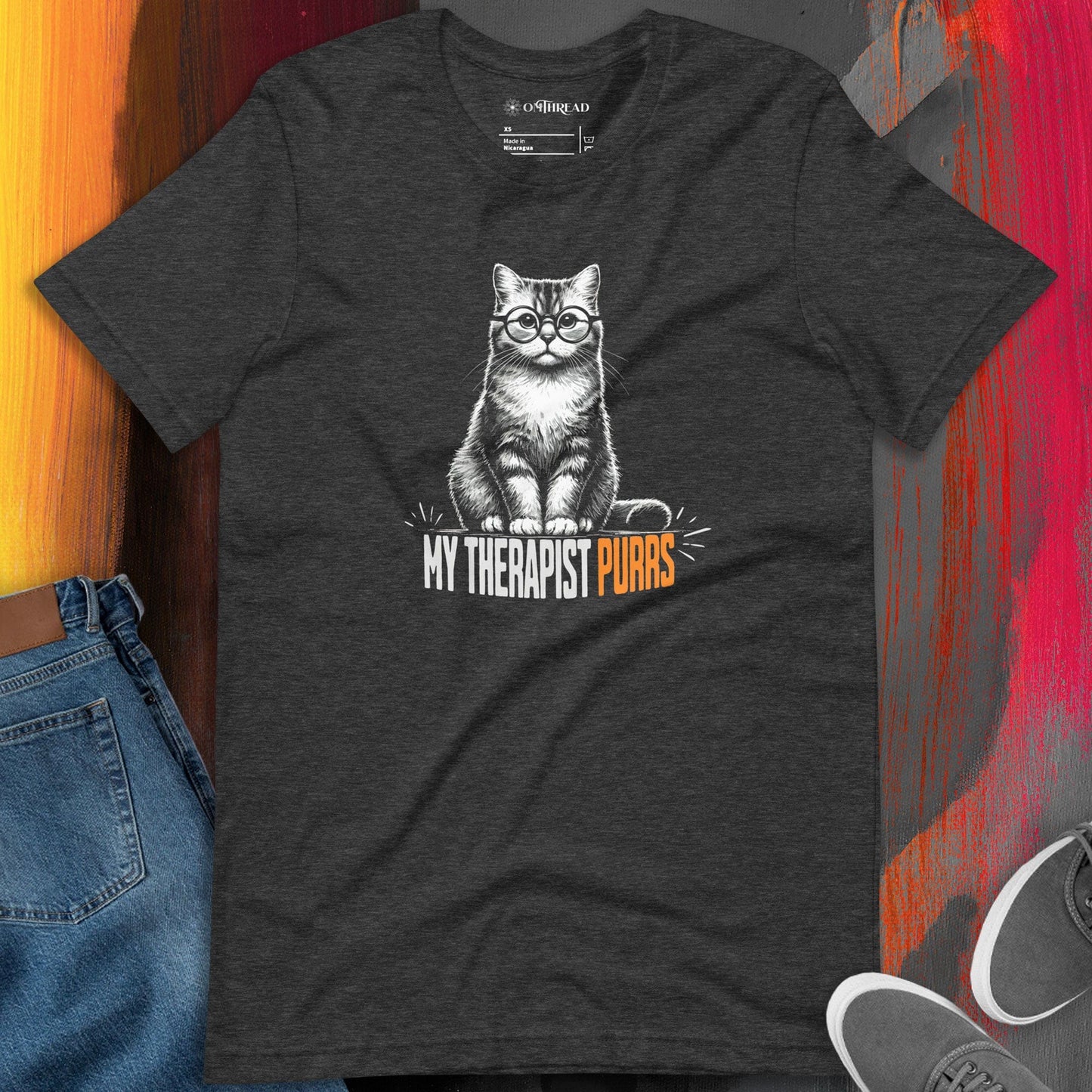 OMTHREAD dark gray t-shirt featuring an illustrated cat wearing round glasses, sitting upright with a calm expression. Below the cat, bold text reads "My Therapist Purrs," with "Purrs" highlighted in orange.