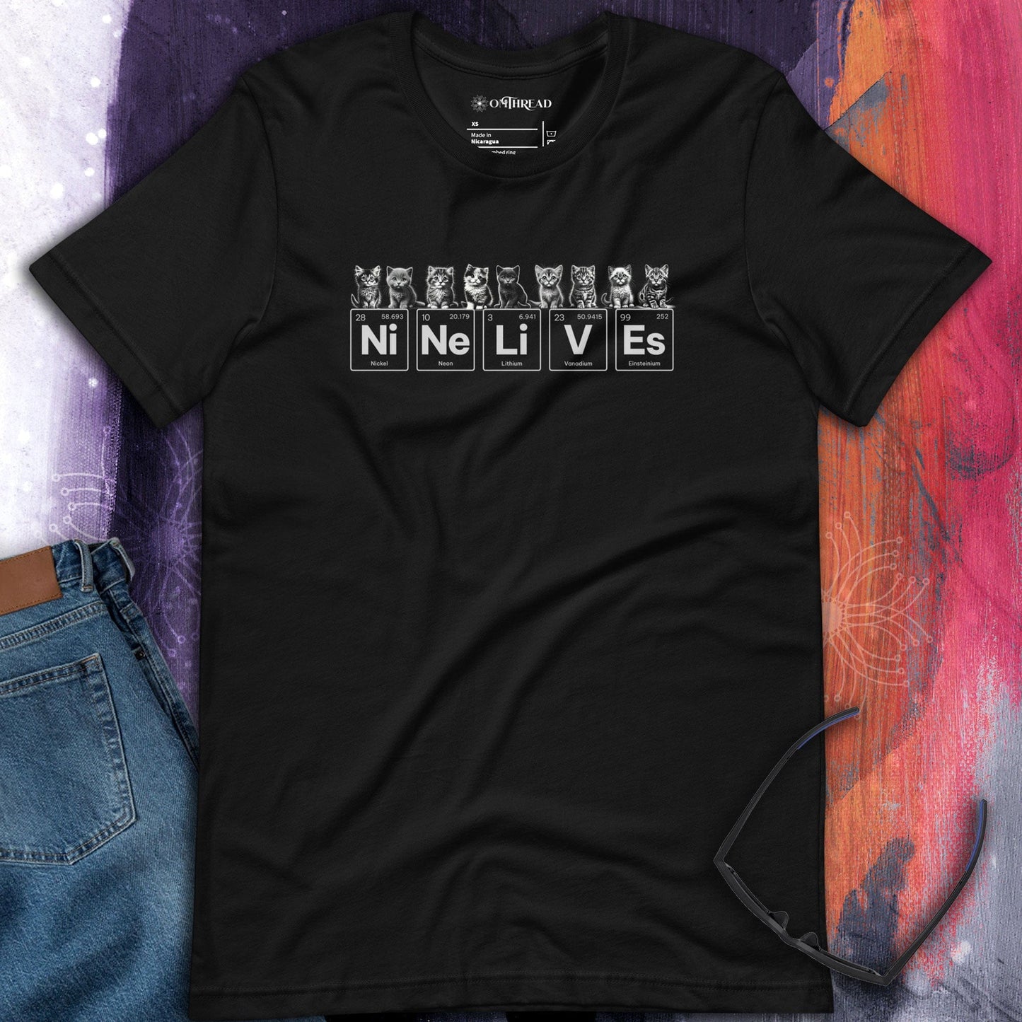 OMThread black t-shirt featuring nine illustrated kittens with playful expressions, sitting above periodic table elements that spell 'Ni Ne Li V Es' (Nickel, Neon, Lithium, Vanadium, Einsteinium), combining science and feline humor.