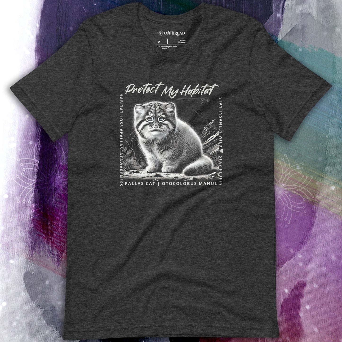 OMThread charcoal grey t-shirt featuring a black-and-white illustration of a Pallas cat kitten sitting in its natural habitat. The design includes the phrase "Protect My Habitat" at the top, with additional text on the sides reading "Habitat Loss • Pallas Cat Awareness" and "Stay Insane • Stay Wild • Stay Fluffy." The bottom text reads "Pallas Cat | Otocolobus Manul."