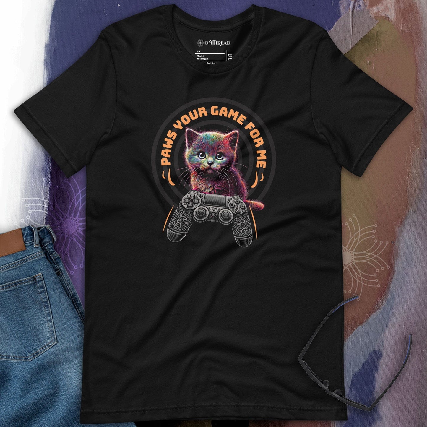 OMTHREAD black t-shirt featuring a neon-style cat holding a video game controller with the phrase "Paws Your Game For Me" in bold orange text. The design blends vibrant colors with a futuristic gamer aesthetic.