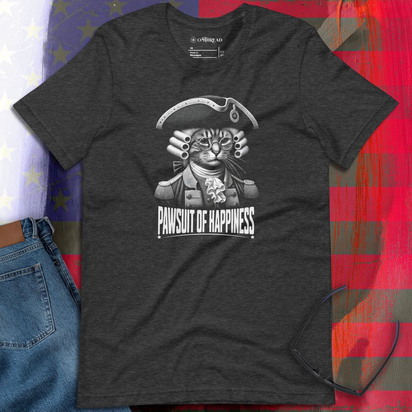 OMTHREAD dark gray t-shirt featuring a detailed illustration of a cat dressed as a founding father in colonial attire, wearing a tricorn hat and glasses. Below, the phrase "Pawsuit of Happiness" is displayed in bold lettering.
