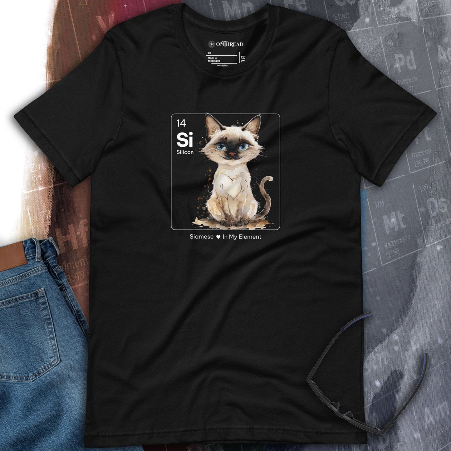 OMThread black t-shirt from featuring a watercolor-style Siamese cat inside a periodic table-inspired square labeled "Si" for Silicon. Below, the text reads "Siamese In My Element." The design combines science and feline charm.