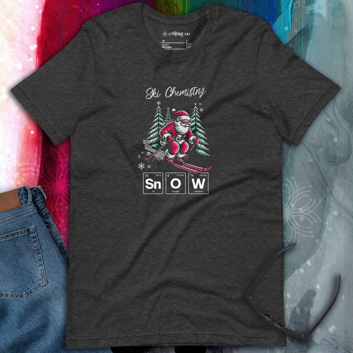OMThread dark gray t-shirt featuring a Santa Claus skiing with a chemistry-themed periodic table design spelling "SNOW," surrounded by pine trees and snowflakes. Holiday-themed science and skiing graphic printed on a soft cotton blend fabric.