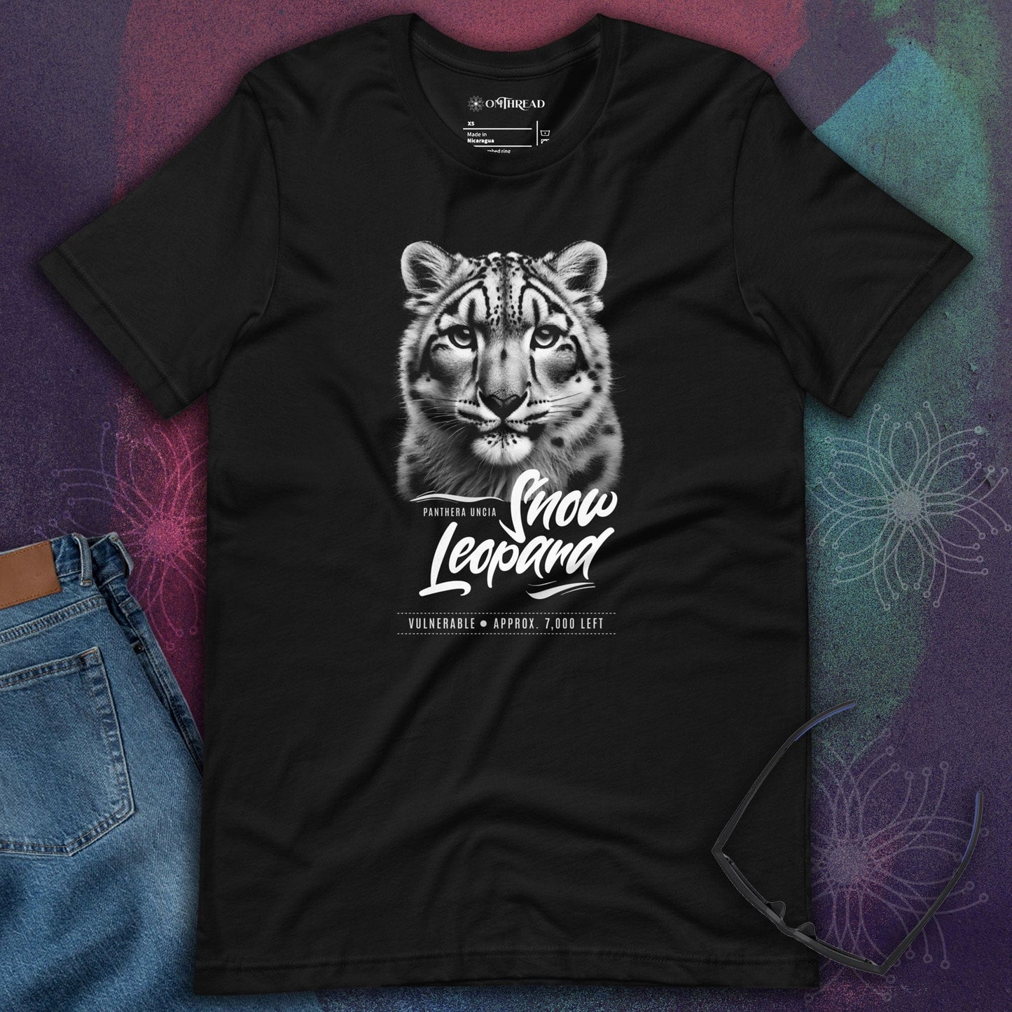 OMThread black t-shirt featuring a detailed black and white illustration of a snow leopard’s face with "Snow Leopard" text below. The design highlights conservation awareness for Panthera uncia. The shirt is laid flat on a textured background with sunglasses placed beside it.
