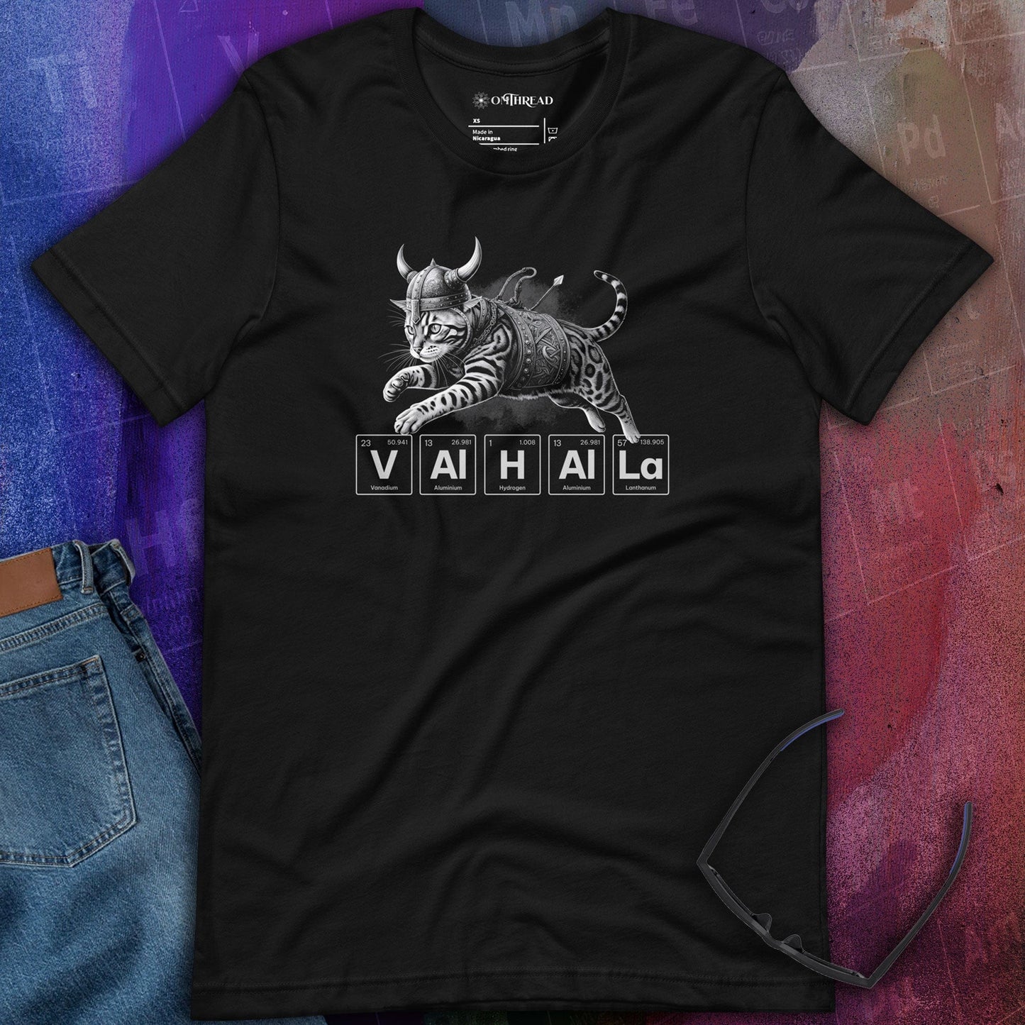 OMTHREAD black t-shirt featuring a Viking-style armored cat in mid-leap. Below the cat, the word "Valhalla" is spelled using periodic table elements, including vanadium, aluminum, hydrogen, and lanthanum.