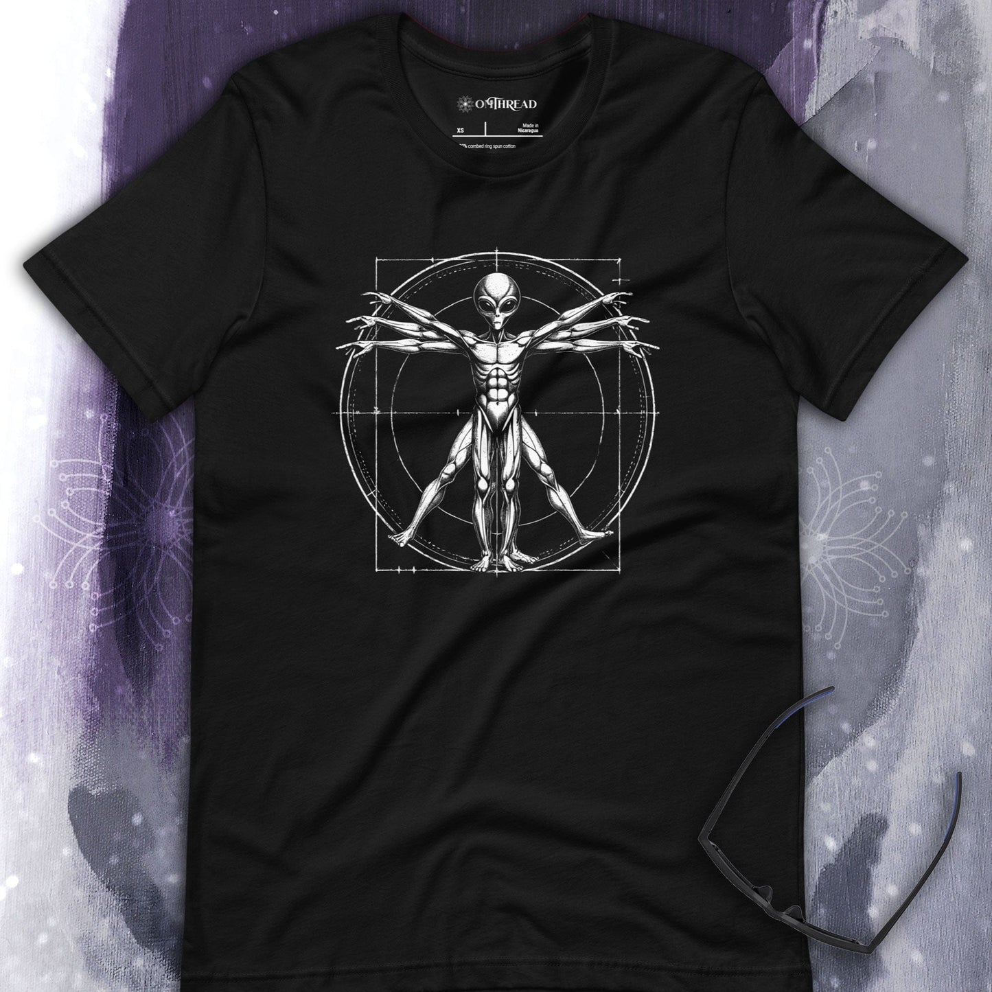 OMTHREAD Vitruvian Alien Tee black t-shirt featuring an alien figure inspired by Da Vinci's Vitruvian Man with detailed anatomy, extended limbs, and a geometric framework of circles and squares.