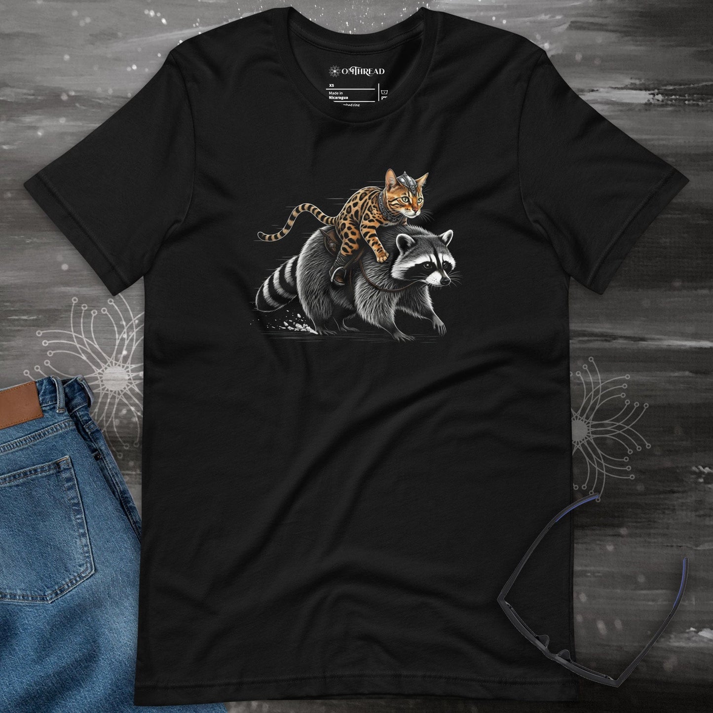 OMTHREAD black t-shirt featuring a Viking warrior cat with Nordic markings riding a determined raccoon. The feline has a fierce expression, gripping the raccoon's fur, while the raccoon strides forward with focus.