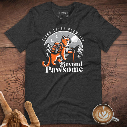 Dark gray t-shirt featuring an adventurous orange cat with a backpack and hiking stick, standing against a mountain and moonlit night backdrop. Text above reads 'Climb Every Meowntain,' and below it says 'Beyond Pawsome' in bold white letters.