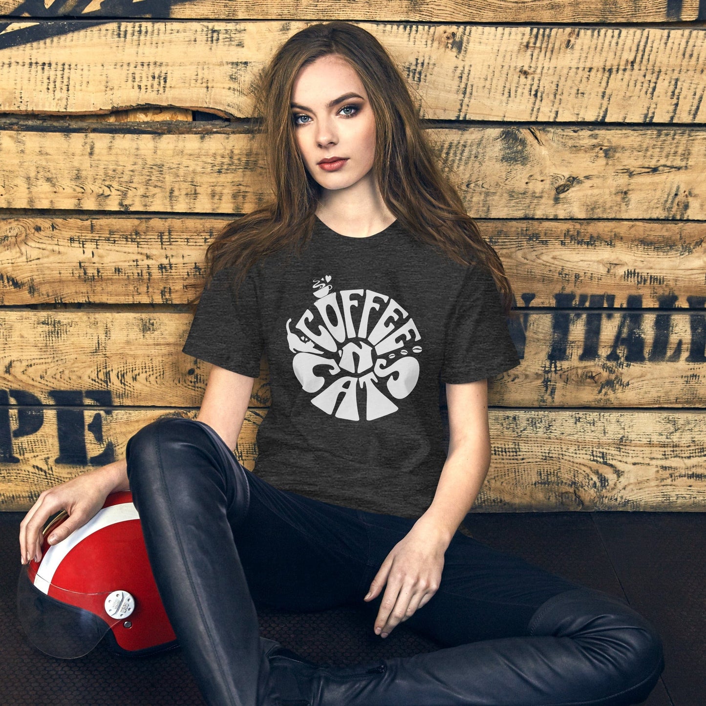 Woman wearing the 'Coffee N' Cats' t-shirt in charcoal gray, seated in a rustic setting with a wooden backdrop, embracing a laid-back, stylish vibe for cat and coffee lovers.