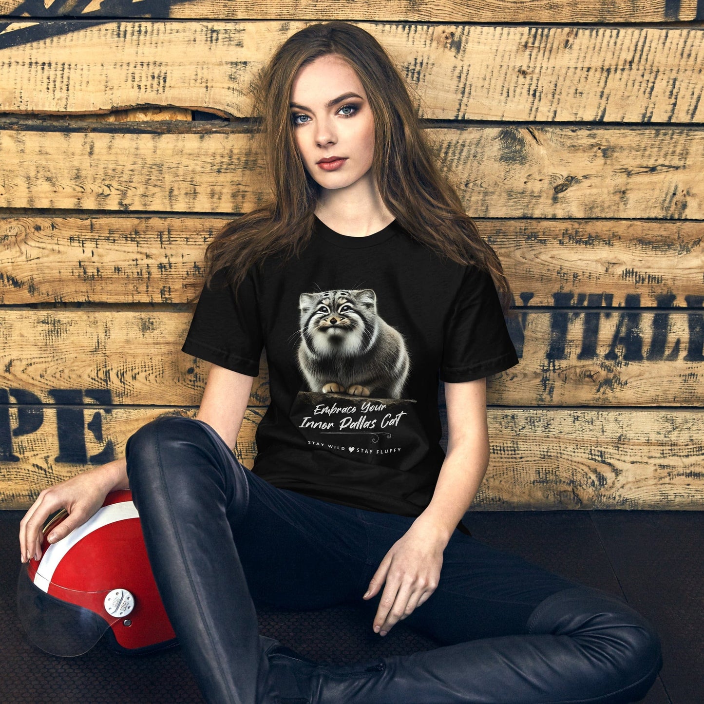 Woman in a black t-shirt featuring a Pallas cat graphic and motivational text, seated against a vintage wood background, blending casual style with nature-inspired design.