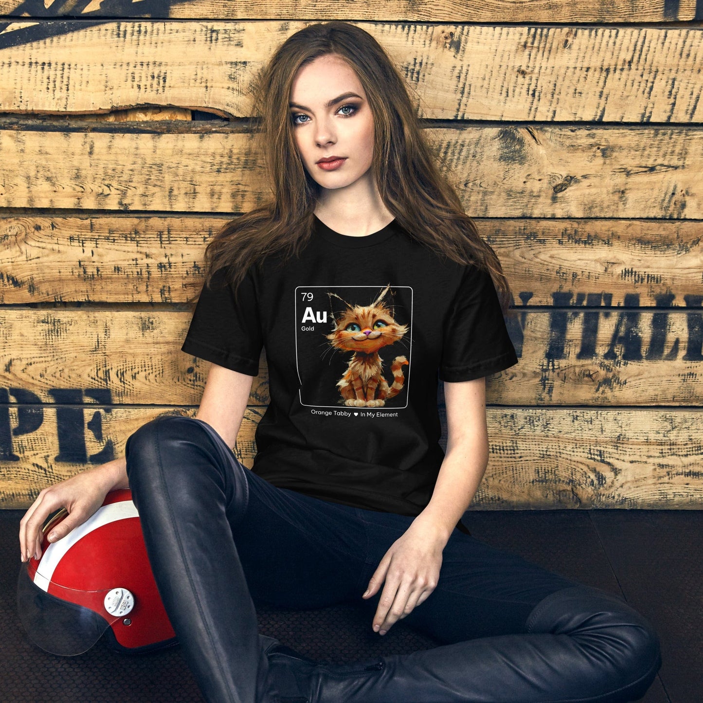 Woman wearing a black t-shirt featuring an orange tabby cat sitting cheerfully inside a square design resembling a periodic table element. The square includes 'Au' for gold, the atomic number 79, and the text 'Orange Tabby In My Element' underneath.