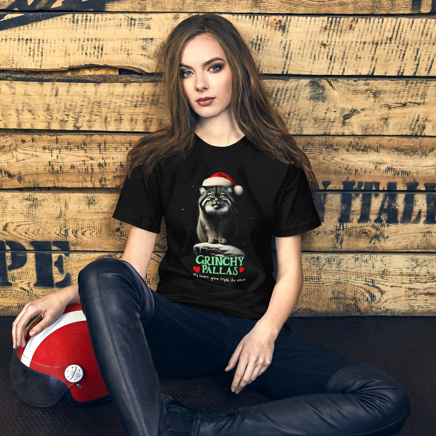 Woman wearing black t-shirt featuring a Pallas cat wearing a Santa hat, sitting on a snowy rock with a grumpy expression. The text below reads 'GRINCHY PALLAS' in green and red, with the phrase 'My heart grew triple its size' underneath.