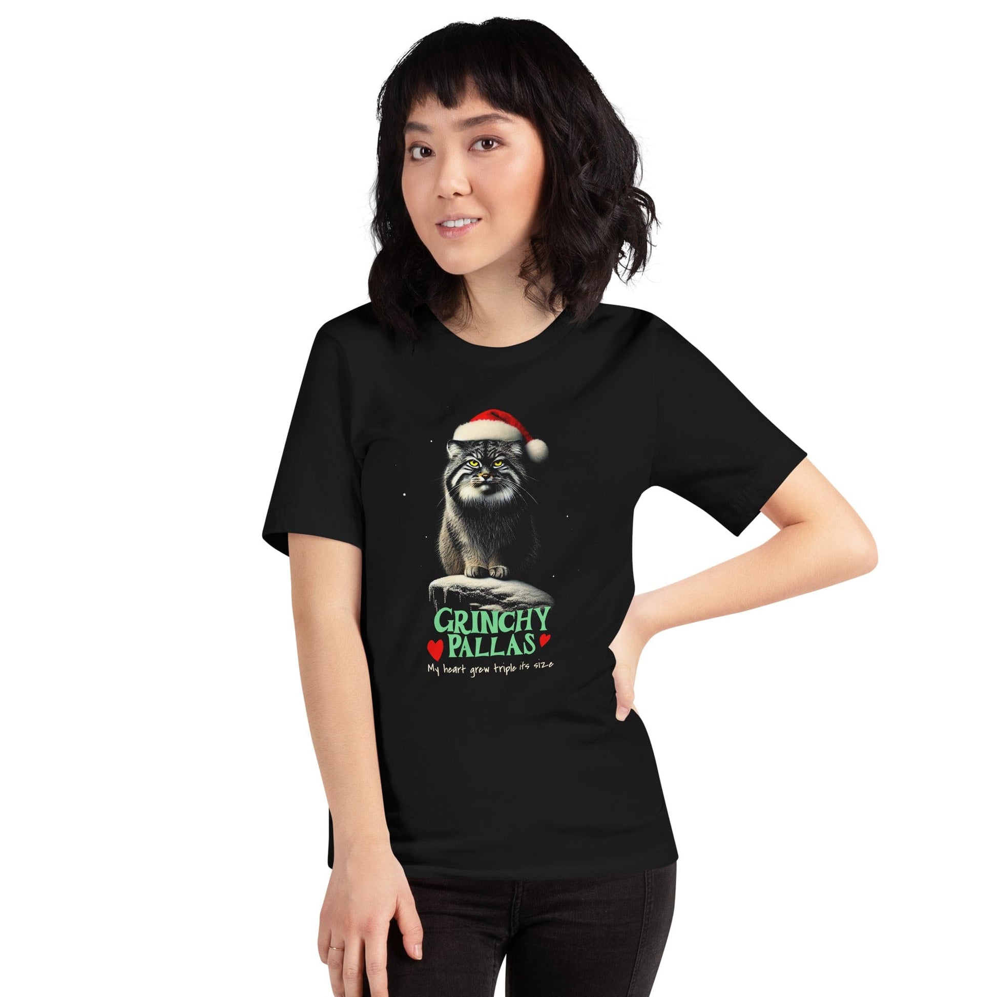 Woman wearing black t-shirt featuring a Pallas cat wearing a Santa hat, sitting on a snowy rock with a grumpy expression. The text below reads 'GRINCHY PALLAS' in green and red, with the phrase 'My heart grew triple its size' underneath.