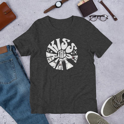 Dark gray t-shirt with a circular, retro-style design that reads 'Kiss the Kitty.' The letters form a sunburst shape with playful illustrations of various cats integrated into the text and surrounding shapes.