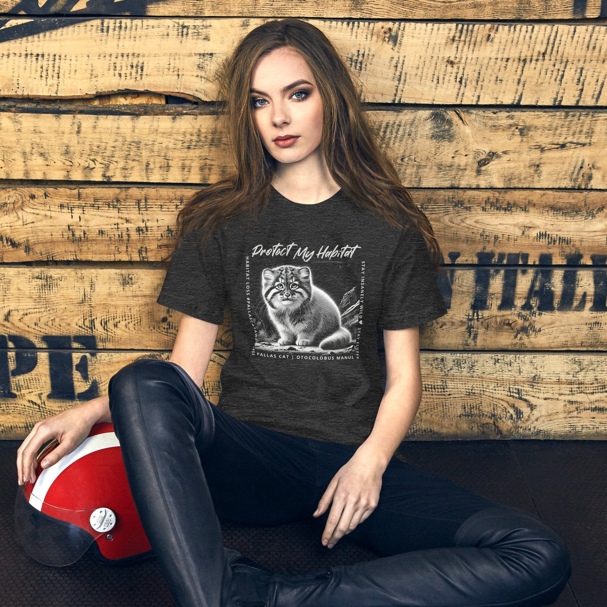 Woman wearing Charcoal grey t-shirt featuring a detailed black-and-white illustration of a Pallas Kitten sitting in its habitat. The design includes the phrase 'Protect My Habitat' at the top, with text on the sides reading 'Habitat Loss Pallas Cat Awareness' and 'Stay Insanely Wild & Stay Fluffy'. The bottom features 'Pallas Cat Otocolobus Manul' in clean typography