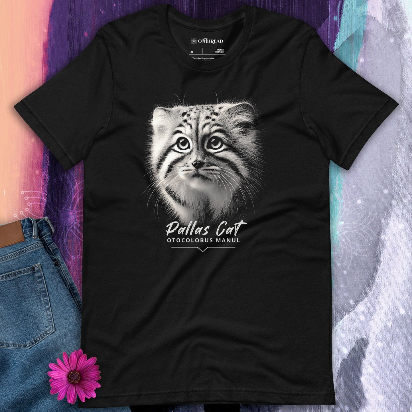 Black t-shirt featuring a close-up, black-and-white illustration of a Pallas kitten's face with striking fur details and intense eyes. Below the image, 'Pallas Cat' is written in elegant white script, followed by 'Otocolobus Manul' in smaller capital letters.