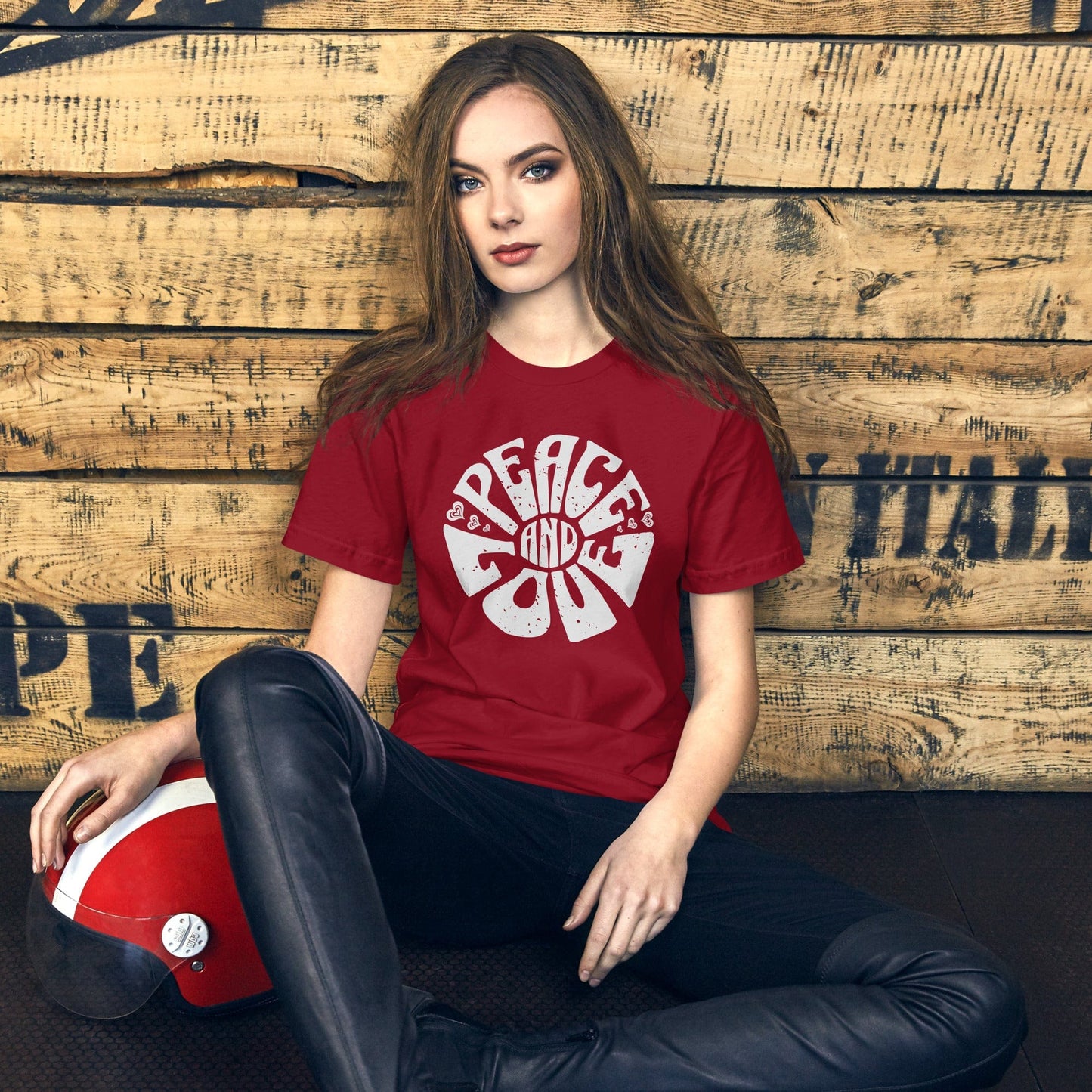 A young woman sitting casually against a wooden wall wears the crimson red "Peace and Love" t-shirt, pairing the vintage graphic with a modern aesthetic. The bold white text stands out on the shirt, creating a striking contrast.