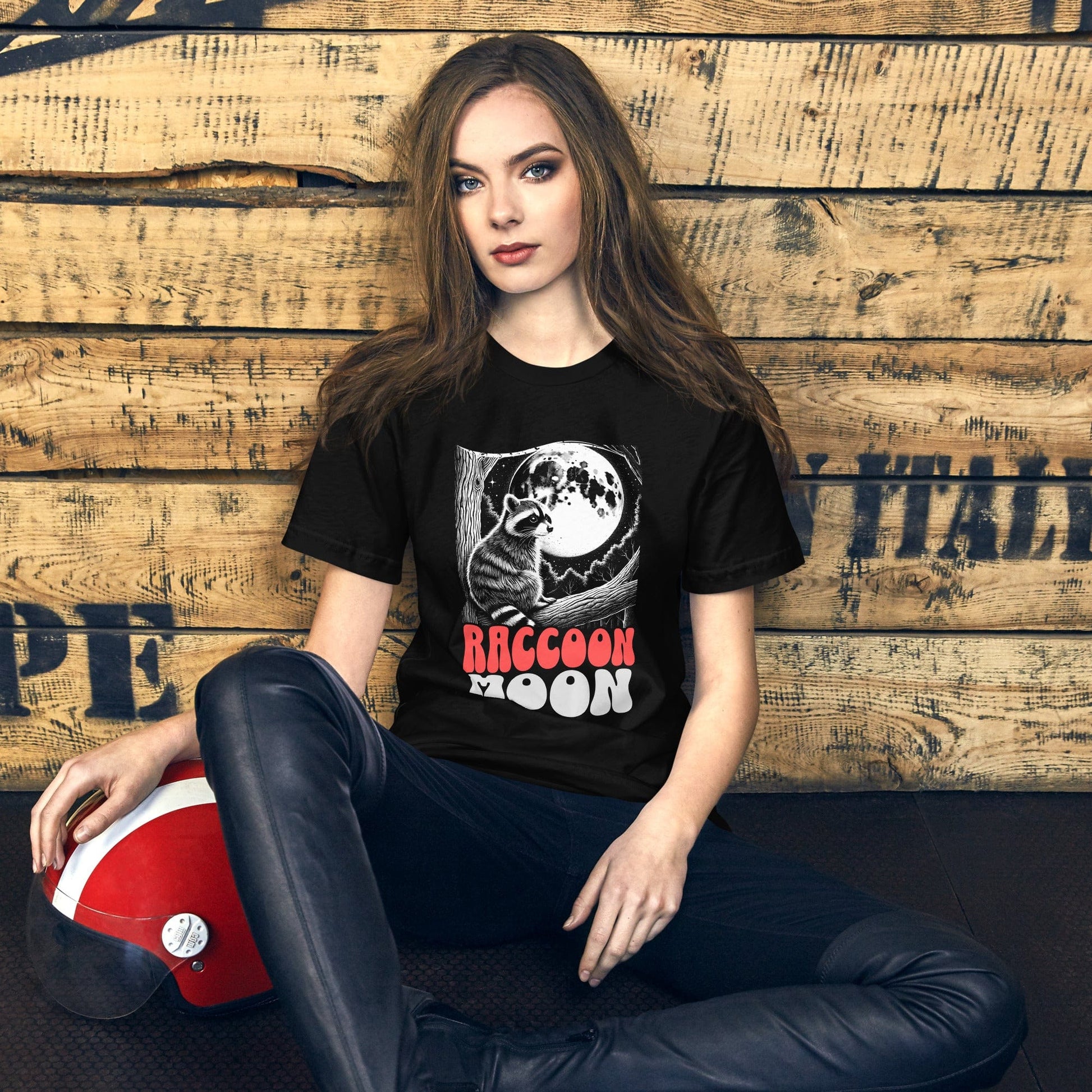 Model wearing the "Raccoon Moon" t-shirt, seated against a rustic wooden background. The design’s bold colors and illustration capture attention, blending a sense of nostalgia with nature-inspired charm.