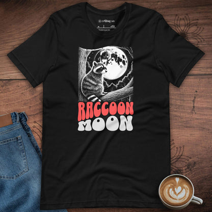 Flat lay of the "Raccoon Moon" black t-shirt on a wooden surface, accompanied by jeans and a latte. The raccoon illustration with the moon is clear and stands out in white and red against the black fabric, giving a serene yet whimsical feel.