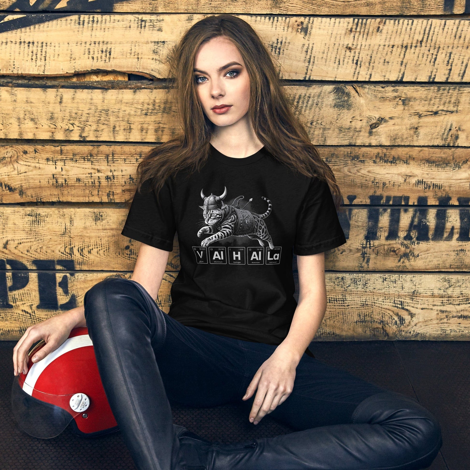 Woman wearing black t-shirt featuring an illustration of a Viking-style armored cat in mid-leap. Below the cat, the word 'VALHALLA' is spelled out using periodic table elements: Vanadium (V), Aluminum (Al), Hydrogen (H), Aluminum (Al), Aluminum (Al), and Lanthanum (La).