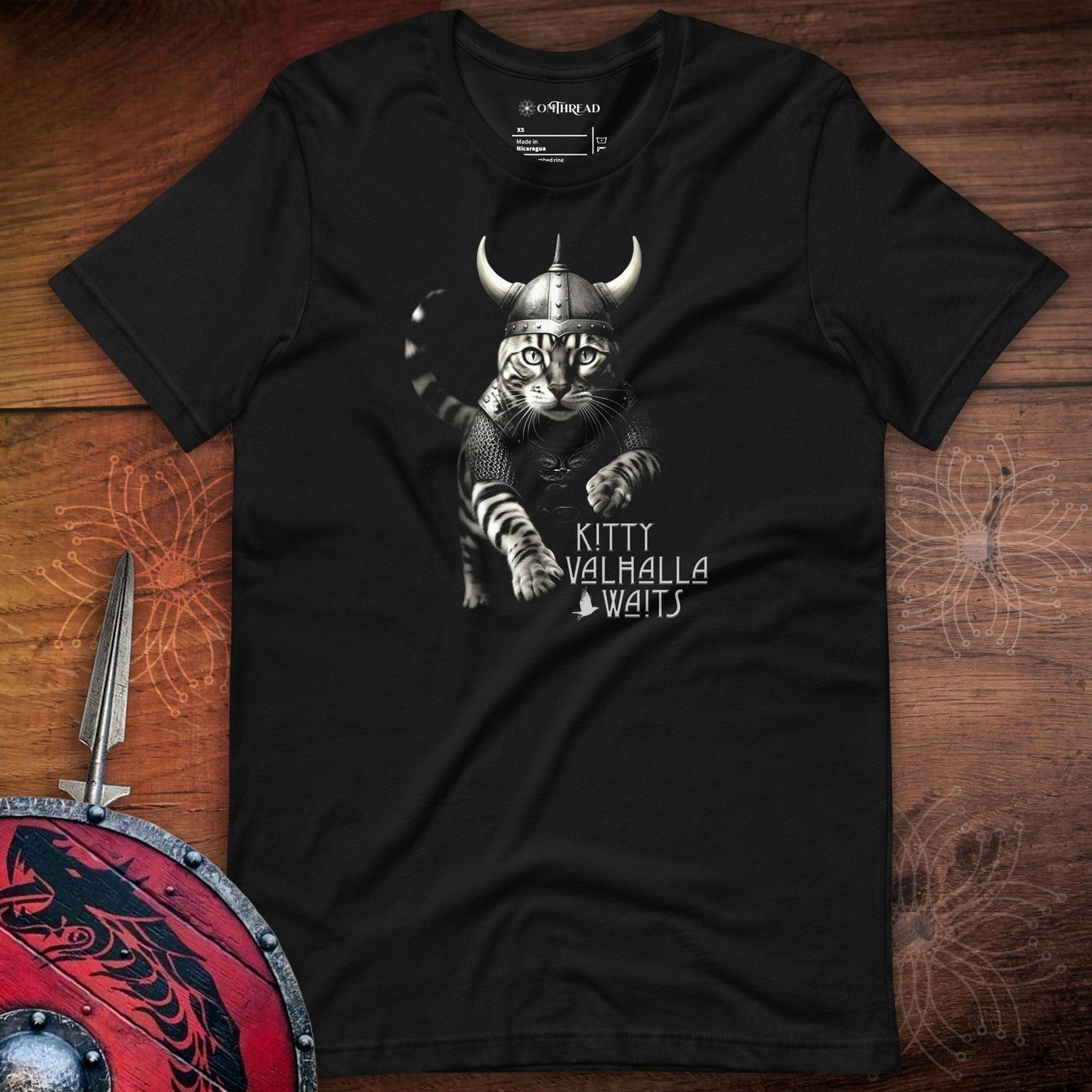 Black t-shirt featuring a Viking-themed design of a fierce bengal cat wearing a horned helmet and armor. The text below reads 'Kitty Valhalla Waits' in bold, stylized font. The background includes Viking elements like a sword and shield.