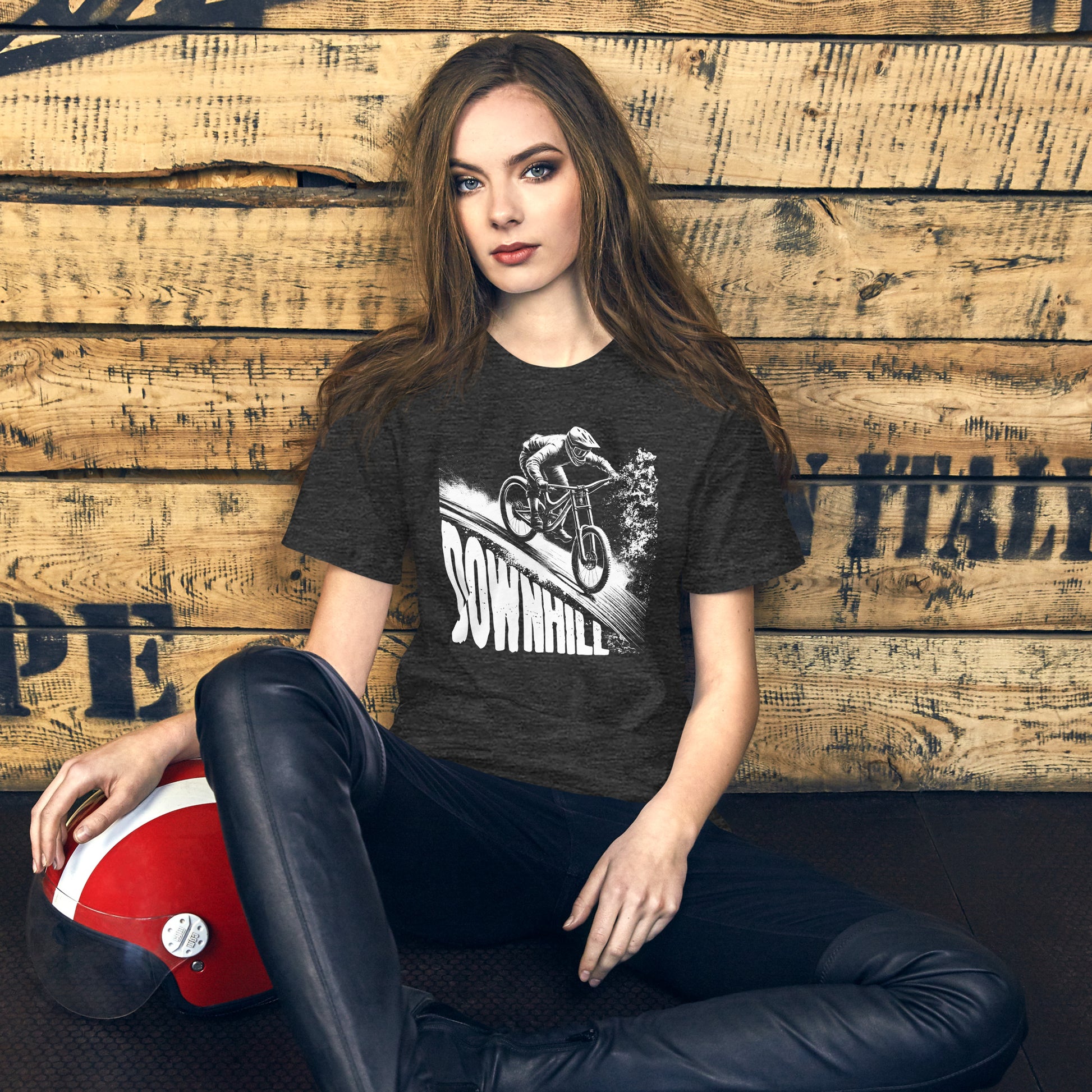 Female model in a rustic setting, wearing a heather gray t-shirt with a bold white 'DOWNHILL' design that features a cyclist racing downhill, emphasizing adventure and outdoor sports.